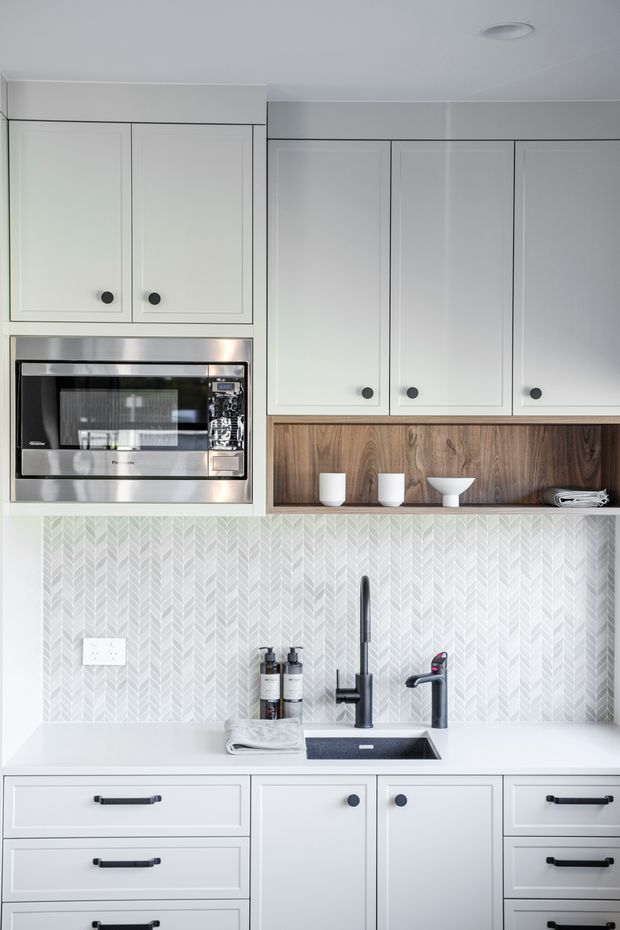 A bright, compact kitchen utility wall with white Shaker-style cabinetry, black round knobs and bar pulls, a stainless-steel microwave, white benchtop, black sink and tapware, and a pale patterned tile splashback. A warm timber open shelf with simple white ceramics provides contrast above the work surface.
