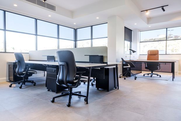 Bright contemporary open-plan office interior with multiple black workstations and ergonomic office chairs arranged around a central desk cluster. Large corner windows provide abundant daylight, while exposed ceiling services, recessed downlights, and track lighting contribute to a clean commercial workspace aesthetic. The floor is finished in large pale grey tiles, with white walls and columns and dark framed glazing.