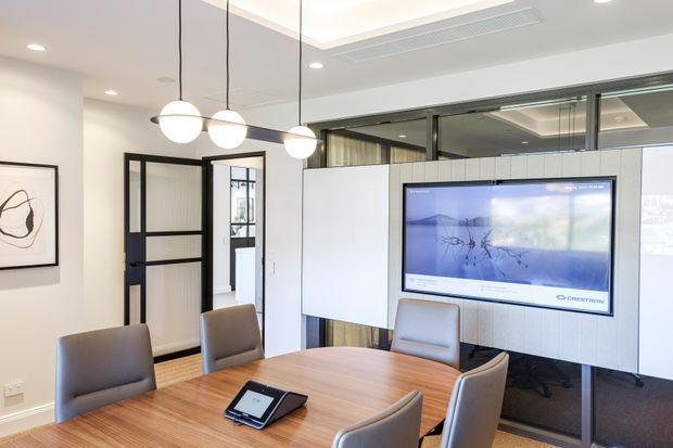 A bright contemporary meeting room or boardroom with a large oval timber conference table, upholstered grey chairs, suspended spherical pendant lights, white built-in wall cabinetry, a wall-mounted display screen, and black-framed glazed partitions and doors. A carpeted area and adjacent glazed workspace are visible beyond the meeting zone.