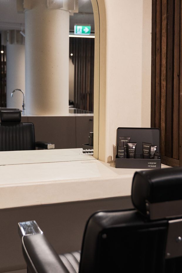 A refined commercial salon or barber workspace viewed from behind a black leather styling chair. A large arched mirror sits above a pale stone or solid-surface counter with integrated basin and tap, reflecting another chair and the interior. Dark timber wall cladding, warm neutral finishes, built-in cabinetry, and displayed grooming products create a polished, contemporary atmosphere.
