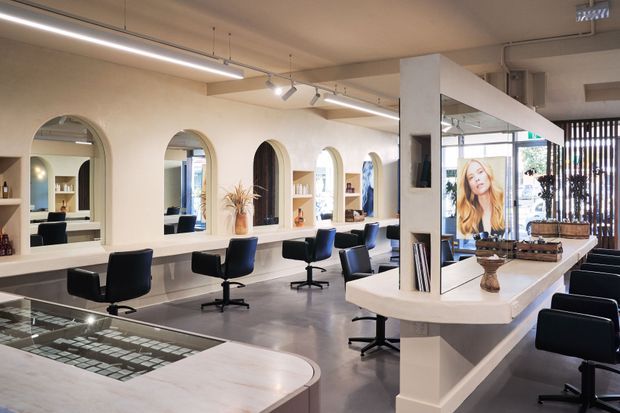 A bright, contemporary hair salon with multiple styling stations, black salon chairs, large arched mirrors, built-in shelving, and a central white island counter. Track and linear lighting illuminate polished concrete-style flooring, while a display screen and vertical timber slats appear near the glazed frontage.