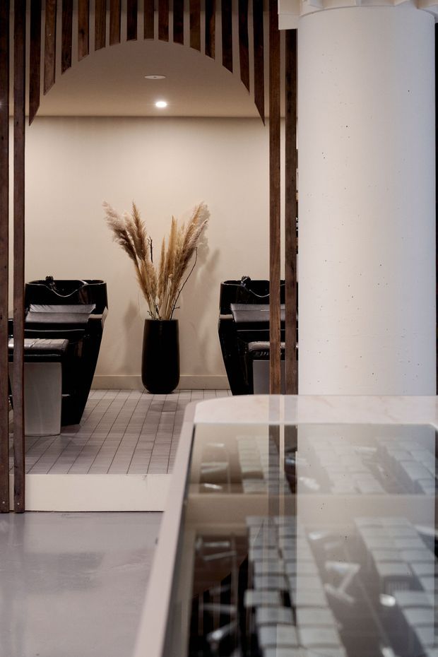 Interior view of a refined salon or beauty studio. Two black shampoo stations with reclining chairs sit against a warm off-white wall, framed by dark vertical timber battens and an arched ceiling detail. A tall black planter with dried pampas grass provides a central decorative feature. Pale square tiled flooring and a glossy reflective counter or glass-topped surface occupy the foreground.