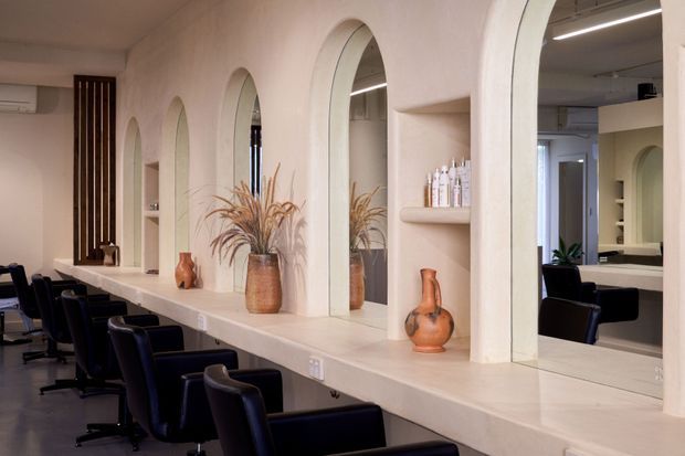 A refined salon interior with a long pale stone or solid-surface styling counter, multiple arched mirrors set into a textured light wall, integrated display niches, and a row of dark salon chairs. Warm ceramic vases and dried botanical arrangements add subdued decorative accents, while ceiling-mounted linear lighting provides functional illumination.