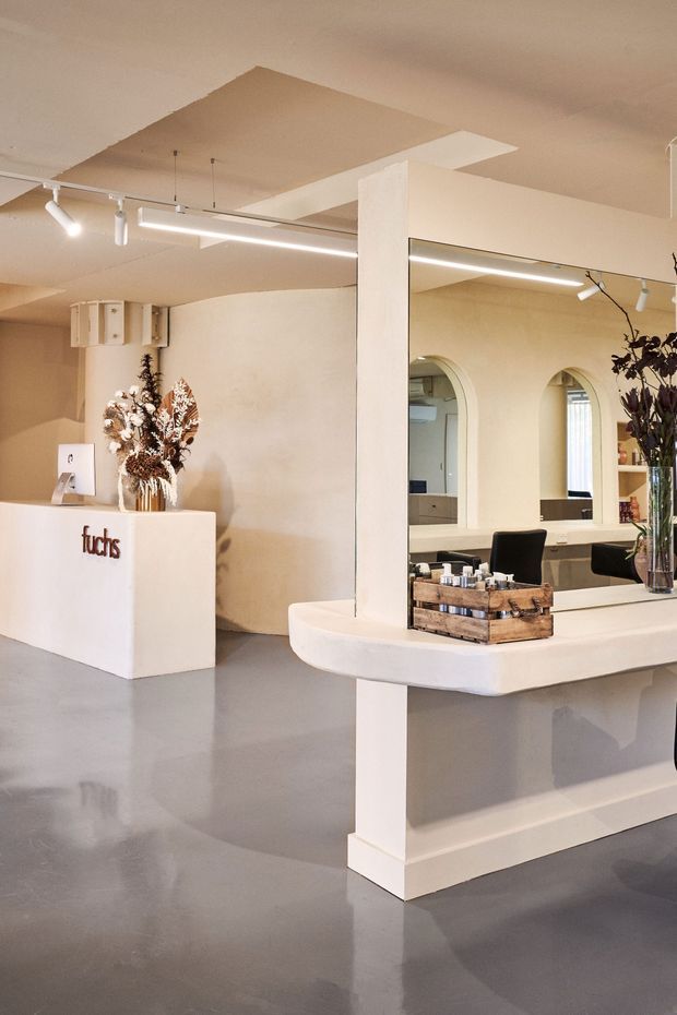 A softly lit contemporary salon or beauty studio interior with a cream-toned reception counter, large wall mirror, arched openings, grey polished floor, white structural columns, track lighting, and dried floral arrangements. A curved white counter with a wooden display tray occupies the foreground, while a branded reception desk is visible at left.