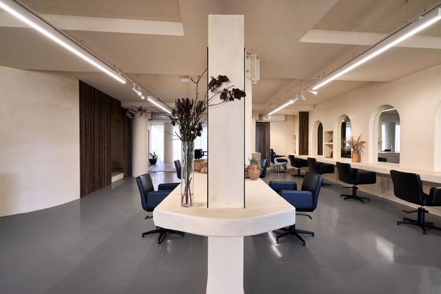 A spacious contemporary hair salon with a central white service island and column, dark salon chairs, long mirrored styling counters, arched mirror openings, grey flooring, and linear LED and track lighting. The palette is predominantly warm white, grey, black, and dark timber, with floral arrangements adding restrained natural accents.