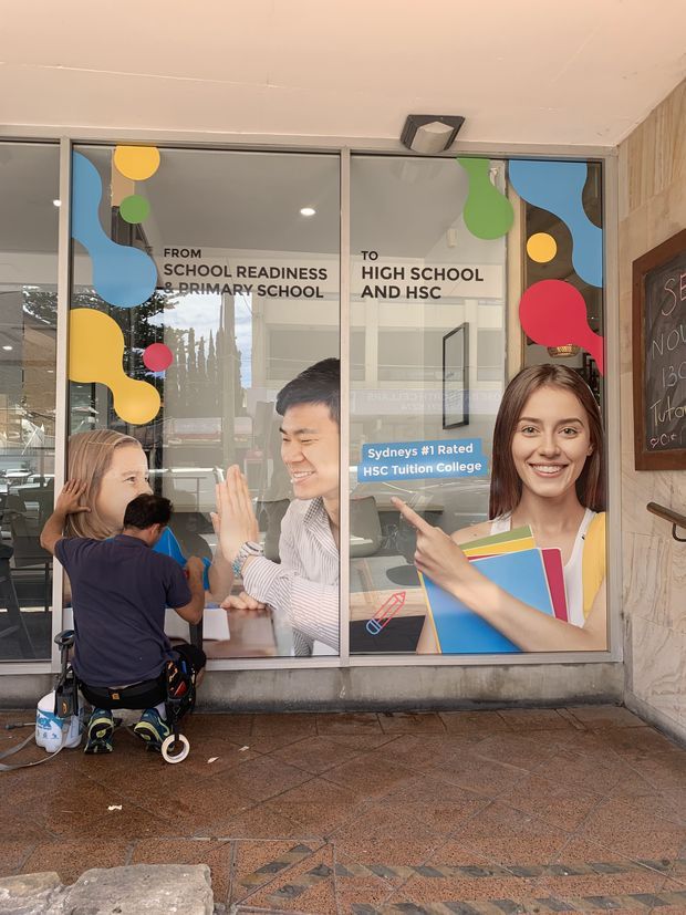 Street-level view of a commercial education facility entrance with large glass window graphics advertising progression from school readiness and primary school to high school and HSC. The glazing features colourful abstract shapes and photographs of students and teachers. A person is seated outside near the entrance, with a chalkboard-style sign at the right and tiled paving in the foreground.