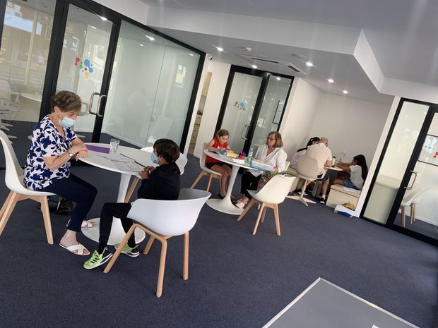 Bright contemporary educational or collaborative workspace with several small round tables, white molded chairs, and people working together. The room has dark carpet flooring, white walls and ceiling, and extensive black-framed glass partitions and doors creating multiple enclosed meeting or activity rooms. The atmosphere is open, functional, and collaborative.
