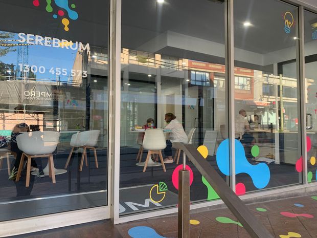 Street-level view through the glazed frontage of a colourful children's learning or therapy centre. The space contains small tables and white molded chairs, with playful multicoloured graphic decals on the windows and pavement. Reflections of the surrounding commercial streetscape are visible in the glazing.