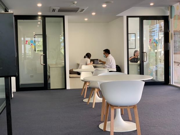 A bright contemporary commercial collaboration or meeting space with dark carpet, white walls and ceiling, recessed downlights, and black-framed glazed meeting-room partitions and doors. White sculptural tables and molded chairs with timber legs occupy the foreground, while two people work at a table in the background. Colourful graphics are visible on the glazing and a large wall graphic appears at right.