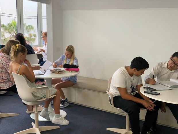 A bright, contemporary shared study or collaborative workspace with several people working at small round tables. The room has white walls, large windows, pale built-in bench seating, white pedestal tables, modern white chairs, and dark carpet flooring. Palm trees are visible outside the glazing.