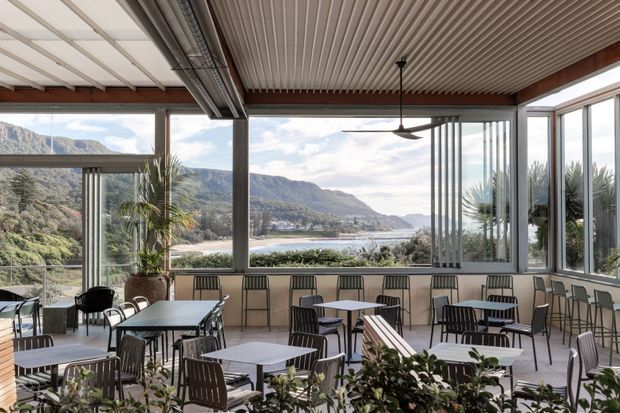 A bright, enclosed terrace-style café or restaurant dining area with extensive floor-to-ceiling glazing overlooking a mountainous coastal or riverside landscape. The space contains numerous small dining tables and metal-framed chairs, a long built-in bench, potted tropical plants, exposed timber beams, corrugated or ribbed ceiling panels, and a suspended ceiling fan. The interior has a relaxed contemporary hospitality feel with strong indoor-outdoor connection.