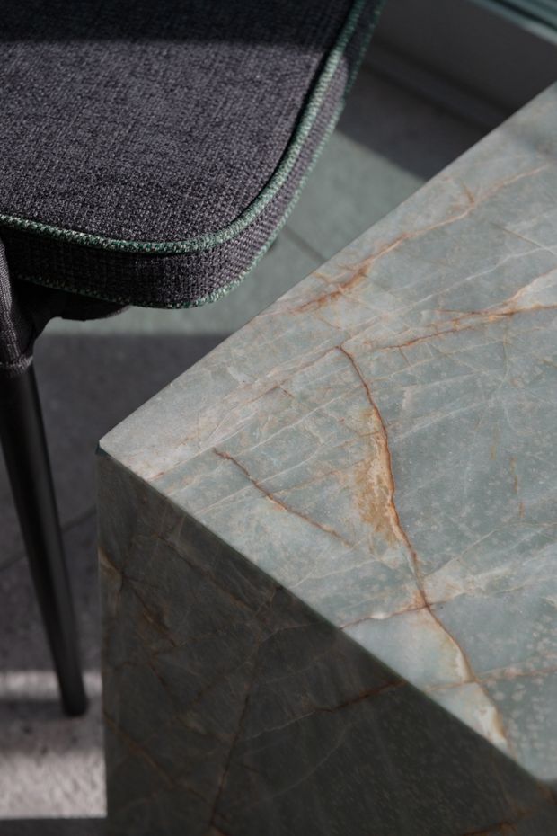 Close-up detail of a dark upholstered chair beside a substantial rectangular stone side table or coffee table. The table has a pale blue-grey natural stone surface with prominent warm rust-coloured veining and matching stone-clad sides, creating a refined material-focused interior vignette.