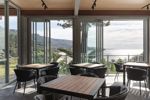 A bright glazed dining or hospitality space with multiple dark-framed timber-topped tables and woven chairs. Full-height folding glass doors open toward a terrace and expansive coastal views, with mountains visible to the side. The interior has a warm timber ceiling, exposed structural beams, concrete-toned flooring, and track lighting.