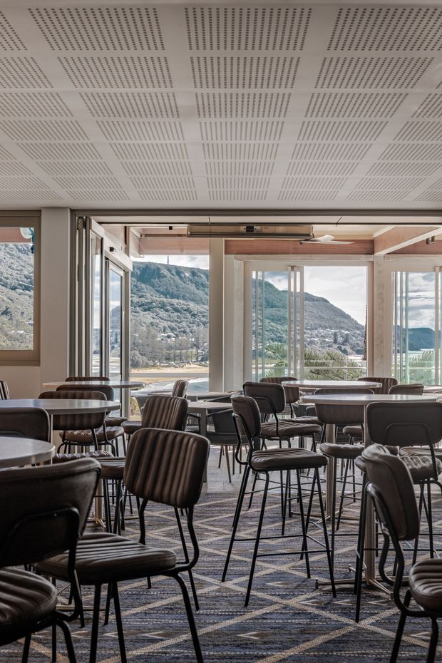 A contemporary commercial dining or lounge space with numerous dark upholstered chairs and small round tables arranged across a patterned carpet. Full-height glazing and open sliding doors provide expansive views toward a mountainous waterfront landscape. The ceiling features a white perforated acoustic panel system, while exposed timber-toned ceiling elements and light structural framing add warmth to the otherwise neutral interior.
