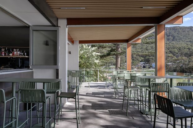 An open-air covered terrace dining and bar area with contemporary green outdoor tables and chairs, a compact bar counter at left, timber slatted ceiling, exposed timber posts and beams, and metal-framed glass balustrades. The terrace overlooks a wooded hillside and is brightly lit with strong indoor-outdoor connection.