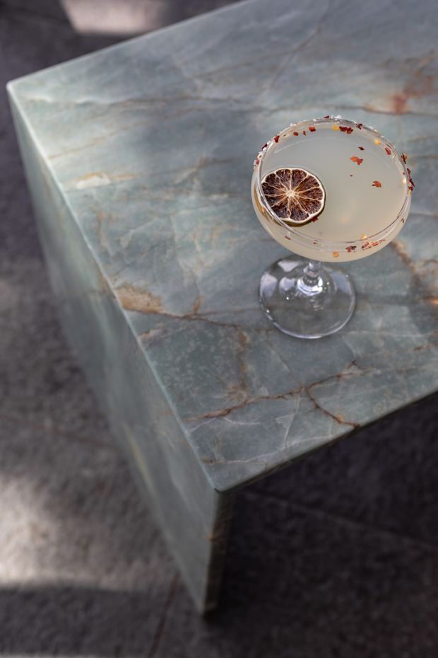 Close-up interior detail of a square coffee table with a thick, irregularly veined blue-grey natural stone slab top and matching stone sides. A coupe glass with a pale cocktail, dried citrus garnish and reddish botanical accents rests on the table. The table stands on a dark textured carpet, creating a moody, refined setting.