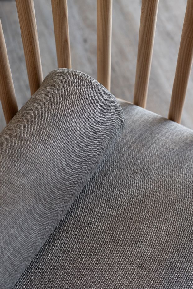 Close-up detail of a grey woven upholstered lounge chair or bench with a cylindrical bolster cushion. Vertical rounded timber slats form the back, with a pale neutral wall visible behind. The image emphasizes tactile textile upholstery, natural timber, and soft neutral tones.