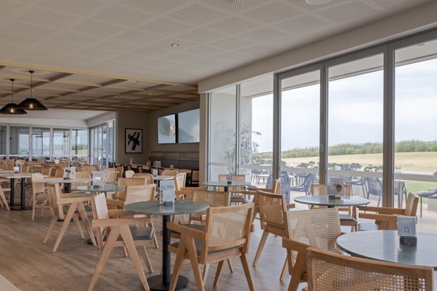 A bright, spacious contemporary restaurant or café dining area with extensive floor-to-ceiling windows overlooking a landscaped outdoor setting. The room features numerous round dining tables, light timber and woven cane chairs, pendant lighting, timber flooring, and a patterned acoustic ceiling. Outdoor terrace seating is visible through the glazing.