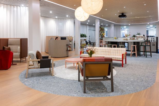 Open-plan contemporary commercial lounge and hospitality-style workspace with multiple seating zones, a reception or service desk, and a kitchenette or bar area. The space features terrazzo-like flooring, light timber flooring, white structural columns, a warm wood-slat ceiling, suspended globe pendant lights, integrated linear lighting, and a muted modern furniture palette.