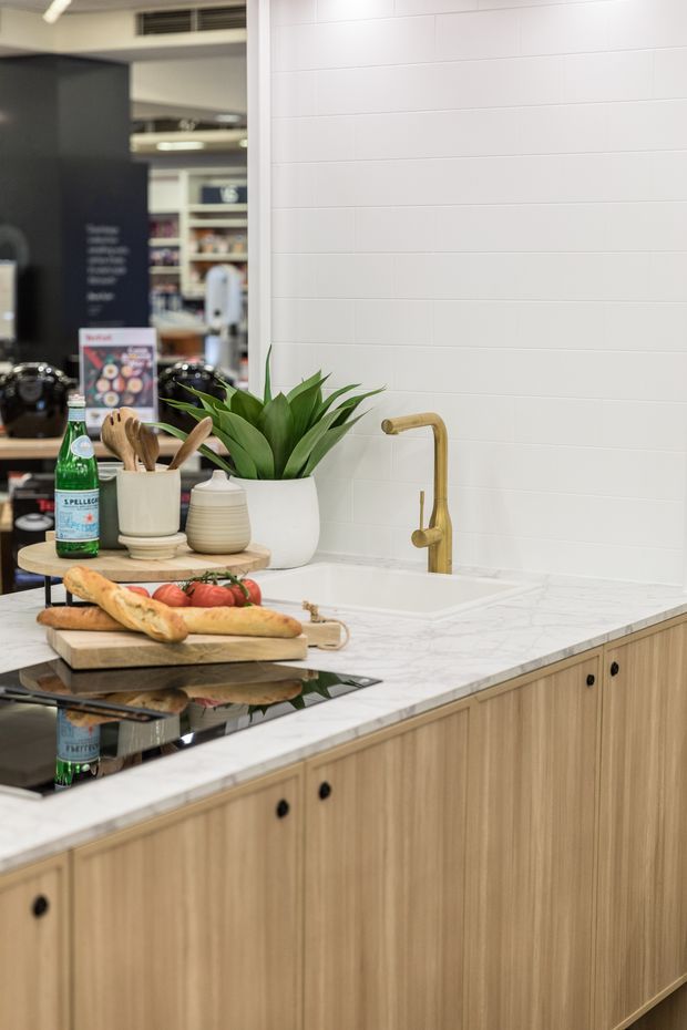 A contemporary kitchen vignette featuring light timber cabinetry with black knobs, a white marble-look benchtop, white horizontal wall tiles, an undermount sink with a brushed brass gooseneck tap, and a black induction cooktop. A wooden serving board with bread and tomatoes, utensils, a potted plant, and countertop items add warmth. Shelving and retail-style displays are visible in the background.