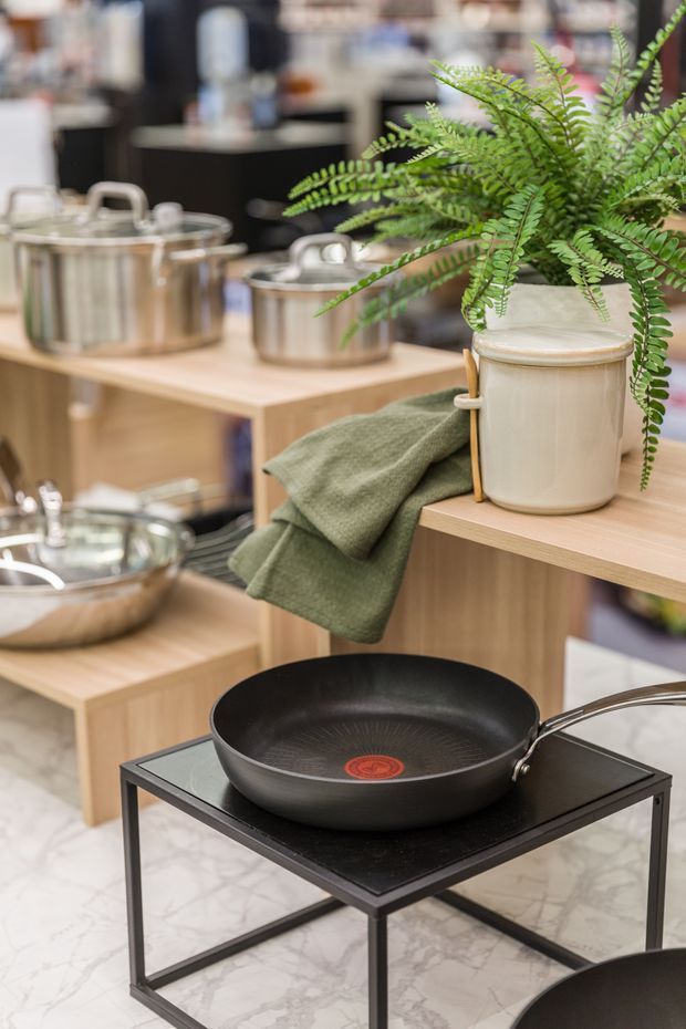 A styled kitchenware display featuring stainless-steel pots, a black frying pan on a small black metal table, a cream ceramic planter with fern, folded green textile, and wooden shelving. The setting appears to be a retail or showroom display, with pale marble-look flooring and warm timber surfaces.