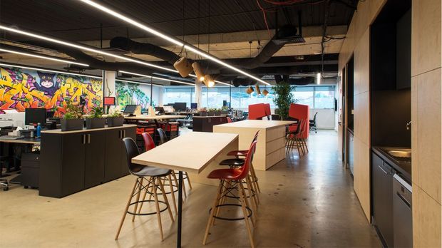 A contemporary open-plan commercial office with shared workstations, long communal white worktables, red and black bar stools, and modern task seating. The space features polished concrete flooring, exposed dark services and ductwork, linear strip lighting, timber wall panelling with integrated kitchenette cabinetry, and colourful graffiti-style wall art.