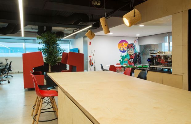 Contemporary commercial office breakout and collaboration area dominated by a large pale timber communal island with red and black stools. The open-plan space includes exposed dark services and ductwork, linear ceiling lighting, woven pendant lights, red acoustic partitions, office workstations, a kitchenette or refreshment counter, and a colourful mural. The setting feels informal, creative, and lively.
