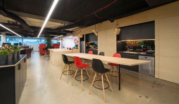 Open-plan contemporary commercial interior with a communal dining or breakout table in the foreground, a kitchenette or refreshment counter at the rear, and work or lounge areas beyond. The space has a polished concrete floor, exposed dark ceiling services, linear pendant lighting, pale timber or wood-look wall panels, and colourful seating and artwork accents.