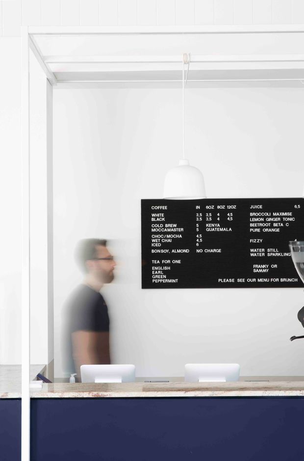 A minimalist café or small commercial coffee bar interior viewed through a large glass opening. The space is predominantly white, with a dark navy service counter, pale stone countertop, white pendant light, simple white stools, and a black menu board mounted on the wall. A blurred staff member is visible behind the counter, reinforcing the café setting. The composition is clean and high-contrast with a restrained contemporary aesthetic.