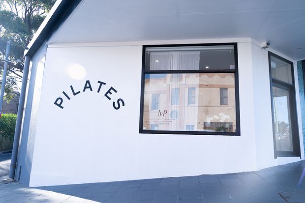 Exterior view of a small Pilates studio with a white rendered facade, bold dark Pilates lettering, black-framed windows and glazed entry door, and a covered flat soffit. Reflections in the windows show surrounding buildings and trees.