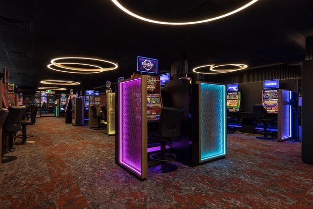 A dark, atmospheric gaming venue lined with electronic slot machines and illuminated gaming terminals. Circular ring pendant lights glow overhead, while vivid magenta, cyan, blue, and amber lighting highlights decorative privacy panels and machines. The space has a patterned carpet and a deep charcoal ceiling, creating a dramatic casino-style interior.