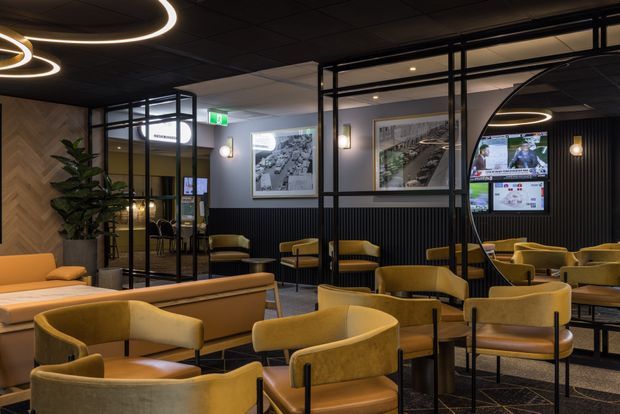A sophisticated contemporary commercial lounge or waiting area with numerous mustard and tan upholstered armchairs and sofas arranged around low tables. The space features black-framed glazed partitions, dark ribbed wall panelling, grey walls, framed artwork, integrated screens, warm feature lighting with circular illuminated pendants, and a timber chevron feature wall with indoor planting.