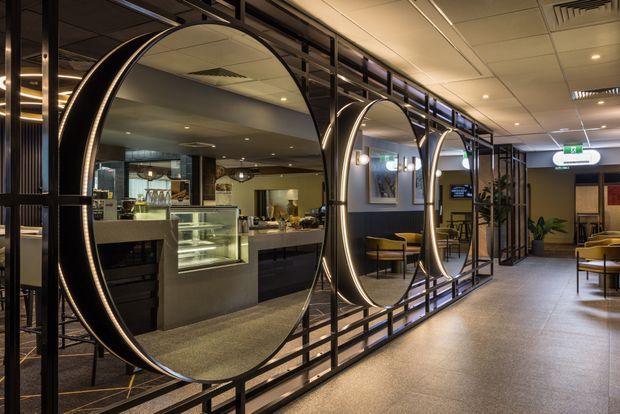 A refined commercial hospitality interior featuring a café or restaurant counter behind a striking black metal partition with large circular mirrors and illuminated curved detailing. The space includes display cabinetry, pendant lighting, café tables and chairs, indoor plants, dark wall finishes, and a tiled ceiling, creating a polished contemporary atmosphere.