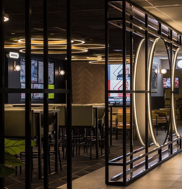 A sophisticated commercial hospitality interior viewed through a black metal framed decorative partition. The space features booth seating and bar-height tables, dark carpet, timber herringbone wall panelling, framed graphics, and dramatic integrated oval and circular LED lighting. The palette is predominantly charcoal, black, warm timber, and muted olive-green, creating a refined, moody restaurant or bar atmosphere.