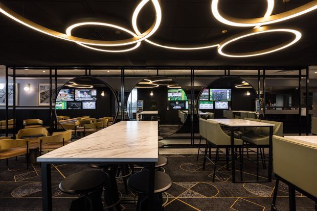 A dark, sophisticated hospitality or entertainment interior with a large marble-topped communal table, bar-height seating, upholstered lounge chairs, patterned carpet, black-framed glazed partitions, and striking circular illuminated pendant lights. Screens and service areas are visible beyond the partitions, suggesting a sports bar or gaming lounge.