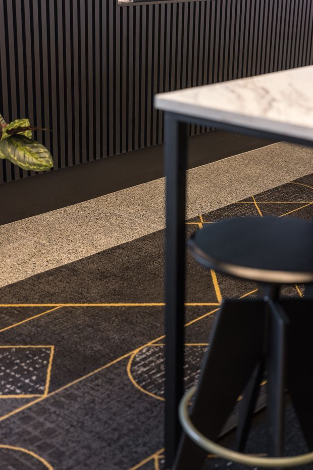 Close-up interior view of a dark, contemporary workspace or hospitality setting. A black-framed table with a pale veined stone or marble-like top and a dark stool with a light metal footrest sit over a charcoal carpet featuring geometric gold linework. The background wall has strong vertical dark timber or slatted panelling, with a small plant adding contrast.