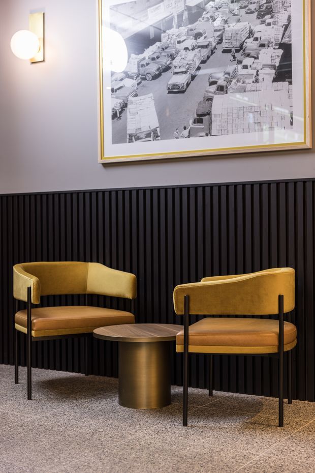 A stylish interior seating vignette with two mustard velvet armchairs arranged around a round bronze-toned side table. The chairs sit on a speckled carpet against a dramatic black vertical slatted wall, with a large framed black-and-white city photograph above and a spherical wall light to the left. The composition has a refined, moody hospitality or commercial-lounge feel.