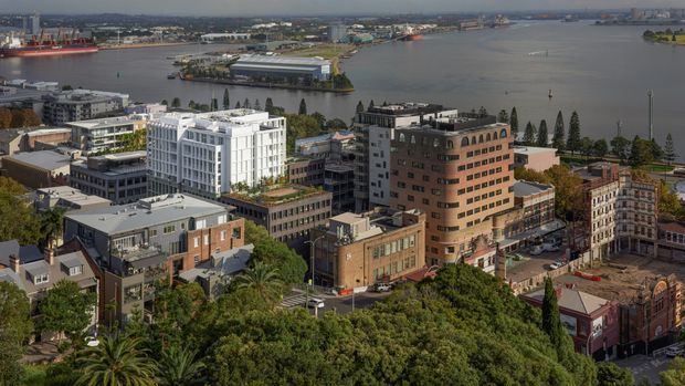 Elevated aerial view of a dense urban waterfront precinct, showing a mix of mid-rise commercial and residential buildings, historic brick architecture, modern white apartment or office buildings, streets, mature trees, and a broad harbour or inlet with port infrastructure in the background.