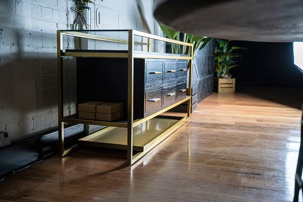 A styled interior featuring a low open shelving and storage console with a slim brass-toned metal frame, dark textured drawer fronts, and wood shelves. The piece stands against a painted concrete-block wall, with polished timber flooring, indoor plants, and soft directional lighting creating a moody, industrial atmosphere.