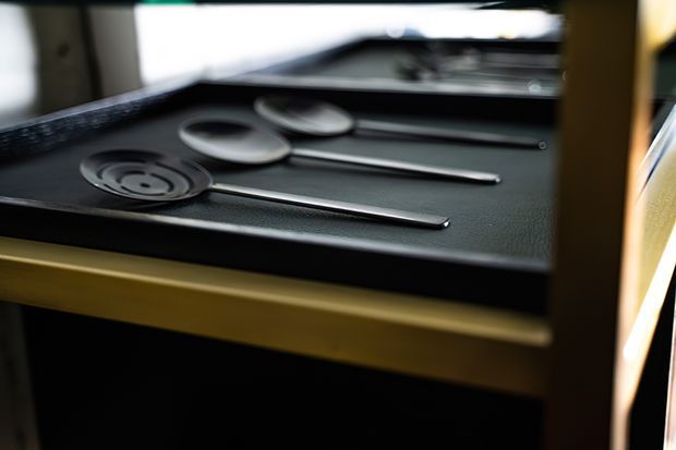 Close-up of a dark textured serving tray or drawer insert holding neatly arranged metal cutlery, including spoons and a fork-like utensil. The tray is framed by warm timber or gold-toned cabinetry, with shallow depth of field and moody directional lighting. The image focuses on storage and presentation details rather than a clearly identifiable room.