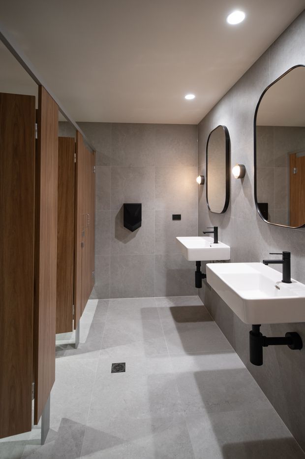 A contemporary commercial-style bathroom or washroom with two wall-mounted white rectangular basins, black wall-mounted taps and exposed plumbing, large rounded-rectangle mirrors, warm wall sconces, and grey large-format tiled walls and floor. Timber-clad cubicle doors line the left side, indicating a shared restroom. Recessed ceiling lights provide even illumination.