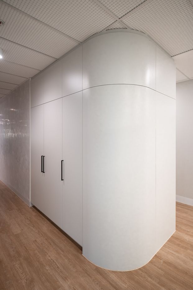 Interior view of a clean, minimalist commercial or institutional space featuring a large curved white built-in cabinetry or partition wall with black vertical handles. The surrounding walls and ceiling are white, with a grid-style suspended ceiling and recessed lighting. Warm wood-look flooring contrasts with the predominantly white fit-out.