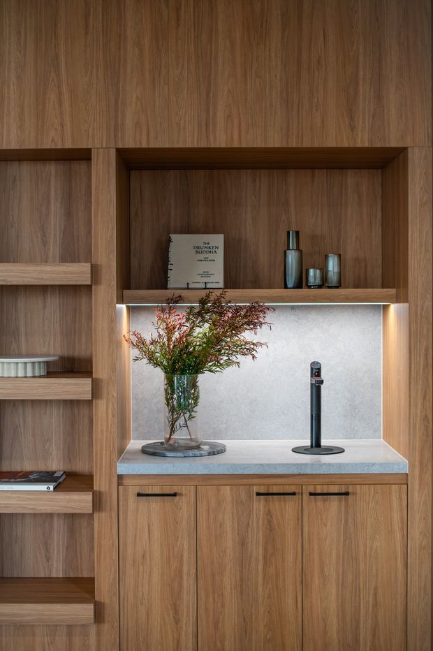 A built-in timber joinery wall featuring open display shelving, a recessed illuminated niche, and lower storage cabinetry. The cabinetry has a warm medium-brown woodgrain finish with dark horizontal handles, while the niche includes a pale stone or engineered-stone benchtop and back panel. Decorative vases, a book, and a floral arrangement are displayed within the joinery.