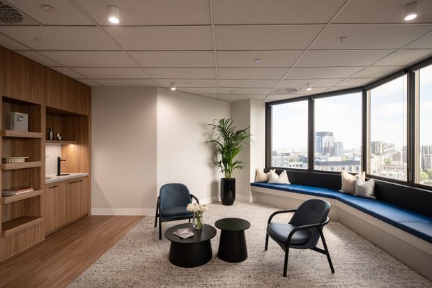 A refined commercial lounge or waiting area with expansive city-view windows, a built-in blue window seat, two dark lounge chairs, round coffee tables, and a tall indoor plant. Warm timber shelving and cabinetry line the left wall, while a suspended acoustic ceiling and recessed lighting create a polished office fit-out.