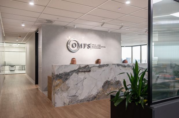 Contemporary commercial reception area with a large veined stone reception desk, branded feature wall, timber-look flooring, glass meeting rooms, and indoor planting. The office has a suspended acoustic tile ceiling, recessed lighting, and a ribbed translucent glass partition.