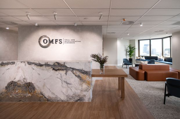 A contemporary medical reception and waiting area with a large veined stone reception desk, timber-look flooring, wood reception table, lounge seating, indoor plants, and expansive city-view windows. A wall sign identifies the practice as OMFS Oral and Maxillofacial Surgeons. The space has a suspended acoustic ceiling with recessed and track lighting.