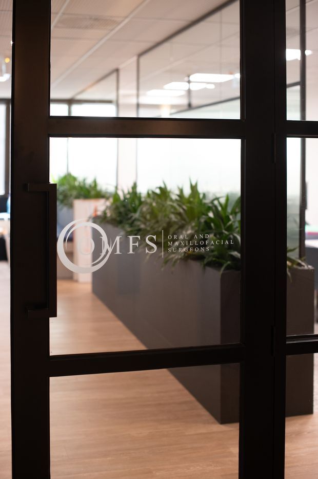 View through a black-framed glazed partition or door into a bright medical or professional office interior. The space beyond has warm timber flooring, large rectangular planters with lush greenery, extensive glazing, and a suspended ceiling with recessed fluorescent lighting. White branding on the glass identifies an oral and maxillofacial surgeons' practice.
