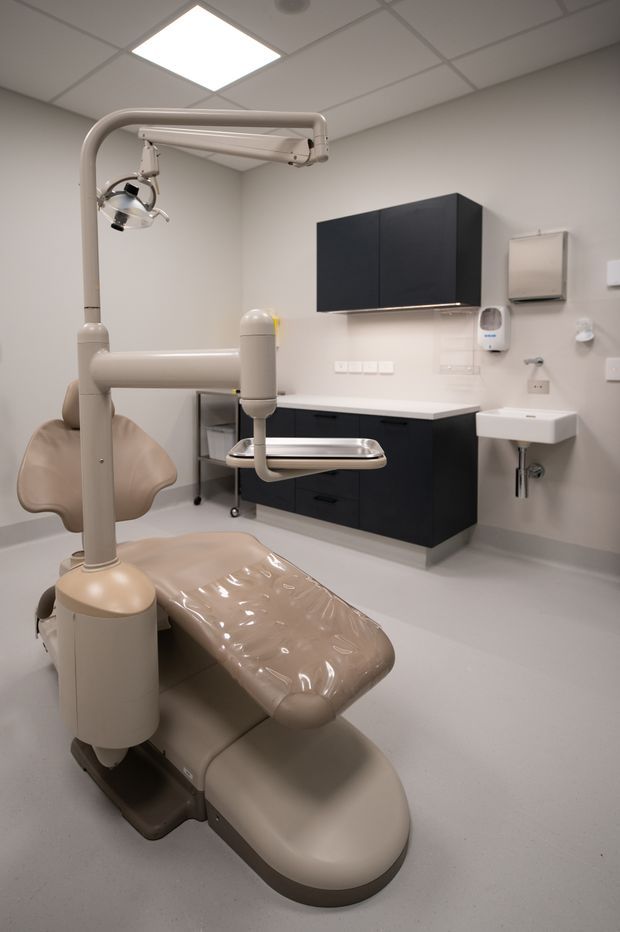 A clean, modern dental treatment room with a large beige dental chair and overhead examination light in the foreground. Dark charcoal wall-mounted cabinetry with a pale benchtop lines the rear wall, alongside a small white hand basin, wall-mounted dispensers, and clinical equipment. The room has smooth light-coloured walls, a pale resilient floor, and a suspended square-grid acoustic ceiling with recessed lighting.