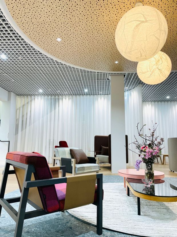 A bright contemporary lounge or reception area with sculptural lounge chairs, round nesting coffee tables, a large rug, sheer floor-to-ceiling curtains, and oversized spherical pendant lights. The space features a distinctive curved perforated timber ceiling panel transitioning into a white exposed grid ceiling, supported by white structural columns. Floral arrangements and layered seating create a refined hospitality-like atmosphere.