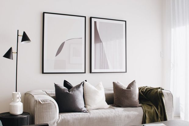 A bright, calm living room with a textured neutral sofa, layered cushions, a draped olive-green throw, black-framed abstract artwork, a minimalist black floor lamp, and sheer curtains filtering daylight.